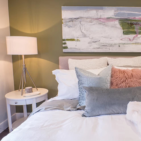 Beautiful bedroom with a sage green feature wall and large piece of art, a pink bed with white and grey bedding and a small round bedside table with a white lamp as featured on Home Network.