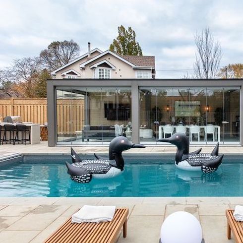 A renovated backyard featured on Home Network’s Backyard Builds featuring large sand patio stones, an inground pool with two blow up loon floaties, an eat at bar, and a covered outdoor kitchen with retractable glass walls by Lumon