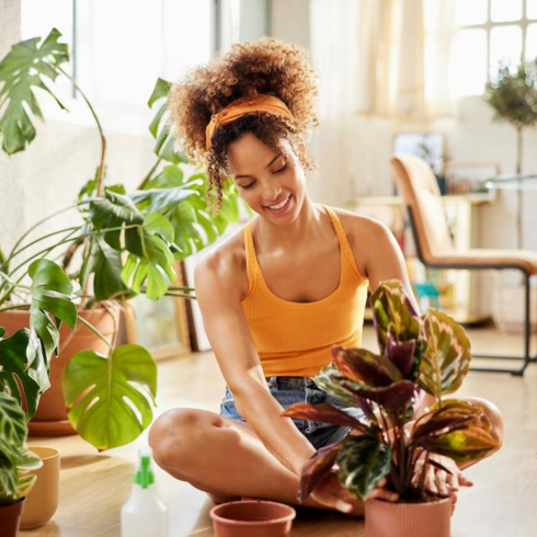 Play Around With Light a young Black woman, with her hair tied back, sitting cross-legged on the floor of her apartment with her plants
